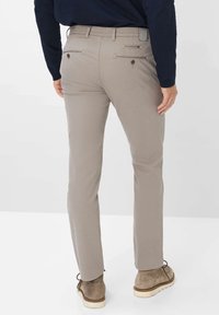 Beige chinos with a straight fit, rear pockets with buttons, smooth fabric texture, and worn with light brown suede shoes.