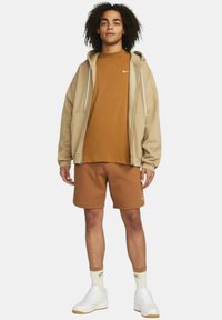 Brun Nike-hoodie över en brun t-shirt, kombinerad med bruna shorts och vita sneakers. Funktioner inkluderar en dragkedja fram och logotypdetaljer.