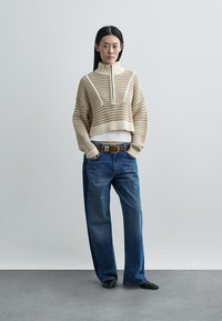 Beżowy krótki sweter z dzianiny z wysokim kołnierzem i teksturowanym wzorem, zestawiony z luźnymi niebieskimi dżinsami i brązowym paskiem.