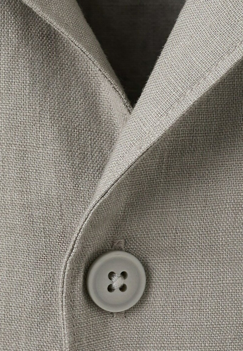 Gros plan sur un blazer en tissu beige tissé avec un col cranté et une fermeture à bouton rond unique.