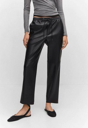 Persoon die zwarte wijde broek van imitatieleer draagt met een trekkoord in de taille, een grijze top met lange mouwen en zwarte platte schoenen met enkelbandjes.