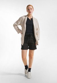 Beiger langer Leinen-Cardigan, schwarzes Kurzarmtop, schwarze Shorts mit Taillenband, weiße Turnschuhe, dunkle Socken. Einfach, legerer Schnitt, ohne Muster.