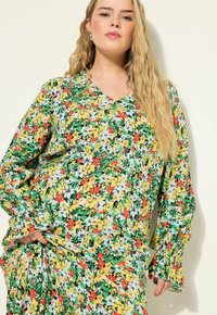 Robe fleurie avec un décolleté en V, présentant un motif vibrant de fleurs jaunes, rouges et blanches sur un fond vert. Manches longues avec des poignets froncés.