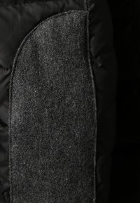 Puderet sort jakke med et grå filt patch på ærmet, som har en blød tekstur og afrundede kanter, der står i kontrast til det glatte basismateriale.