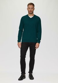 Dunkelgrüner, langärmeliger V-Ausschnitt-Pullover aus weichem Material, kombiniert mit schwarzen Slim-Fit-Jeans und schwarzen Sneakers, vor einem schlichten Hintergrund stehend.