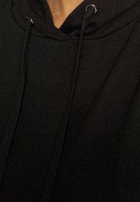 Närbild av en persons nacke i en svart hoodie, med en dragsko med metallspetsar, som visar upp detaljer i texturen på tyget.