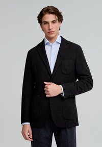Blazer a cuadros negros hecho de tela texturizada, con un solo bolsillo en el pecho, solapas con muesca y cierre de botón, combinado con una camisa de color azul claro.