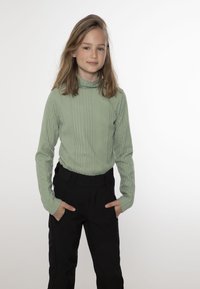 Maglione a collo alto verde a coste, abbinato a pantaloni neri con tasche laterali e una finitura liscia e strutturata.