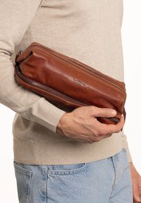 Bruine leren zakje met een cilindervorm, voorzien van een ritssluiting en subtiele, ingebakken branding. Gestructureerd oppervlak, in de hand gehouden.