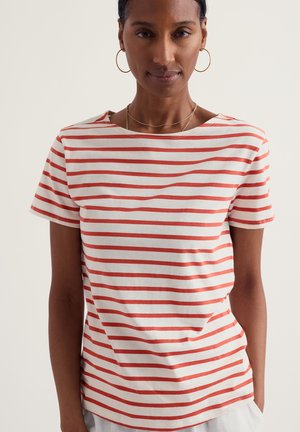 Kobieta w czerwono-białej pasiastnej koszulce z krótkim rękawem, złotych kolczykach kołach i cienkich złotych naszyjnikach na jednolitym tle.