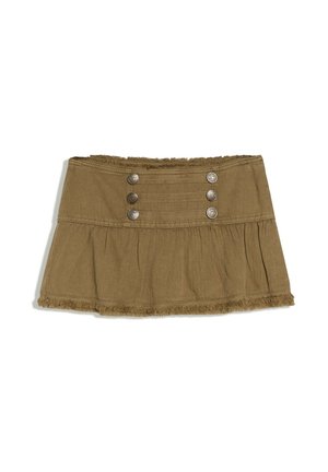 Donna con lunghi capelli scuri che indossa un top a maniche lunghe giallo pallido, una minigonna marrone con bottoni e stivali neri al ginocchio, in piedi davanti a uno sfondo grigio.