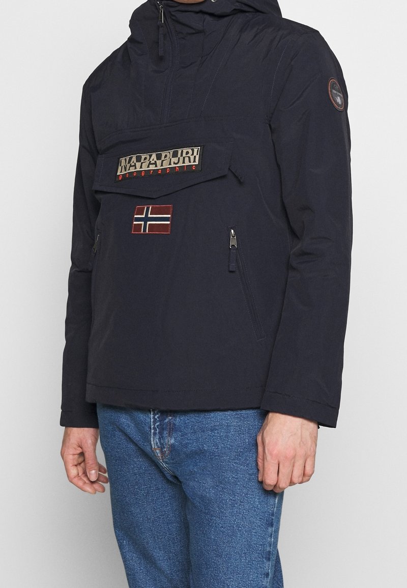 Marineblå pulloverjakke med hætte, stor frontlomme, lynlåslommer på siden og et norsk flag-patch. Lavet af letvægtsmateriale.