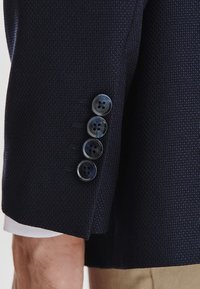 Granatowy teksturowany rękaw marynarki z czterema ciemnymi guzikami o subtelnym połysku. Tkanina ma drobny wzór i gładkie wykończenie.