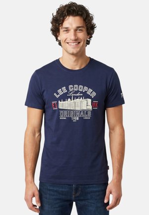 Homme souriant en T-shirt bleu Lee Cooper London Originals avec motif de bâtiment et texte, debout sur fond clair uni.