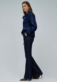 Blusa azul marino con detalle de lazo, combinada con jeans oscuros de campana y zapatos planos marrones de punta, creando un aspecto elegante y coordinado.