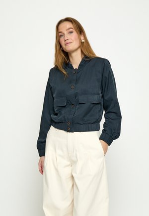 Woman wearing a navy buttoned jacket with flap pockets and white high-waisted wide-leg pants, standing with one hand in pocket.
