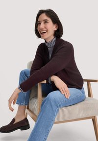 Dunkelviolette gerippte Strickjacke über einem gestreiften Rollkragenpullover, kombiniert mit hellblauen Jeans und braunen Slippern. Person sitzt auf einem Holzstuhl.