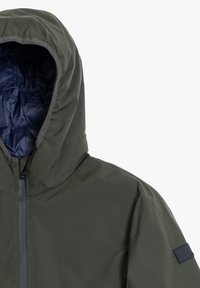 Giacca isolante verde scuro con cappuccio, dotata di un interno blu navy, zip frontale e un accento quadrato blu navy sulla manica.