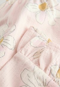 Tela rosa claro con un tejido texturizado, que presenta un patrón floral en blanco y amarillo. Incluye detalles fruncidos y un bolsillo.