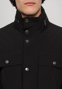 Zwarte jas met een hoge kraag, zilveren hardware, ritssluiting en voorflappen. Gemaakt van gladde, matte stof met een gestructureerd ontwerp.