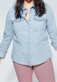 Lichtblauwe denimoverhem met lange mouwen, knoopsluiting aan de voorkant, twee borstzakken en subtiele stikdetails, gedragen met rood-witte geruite broek.