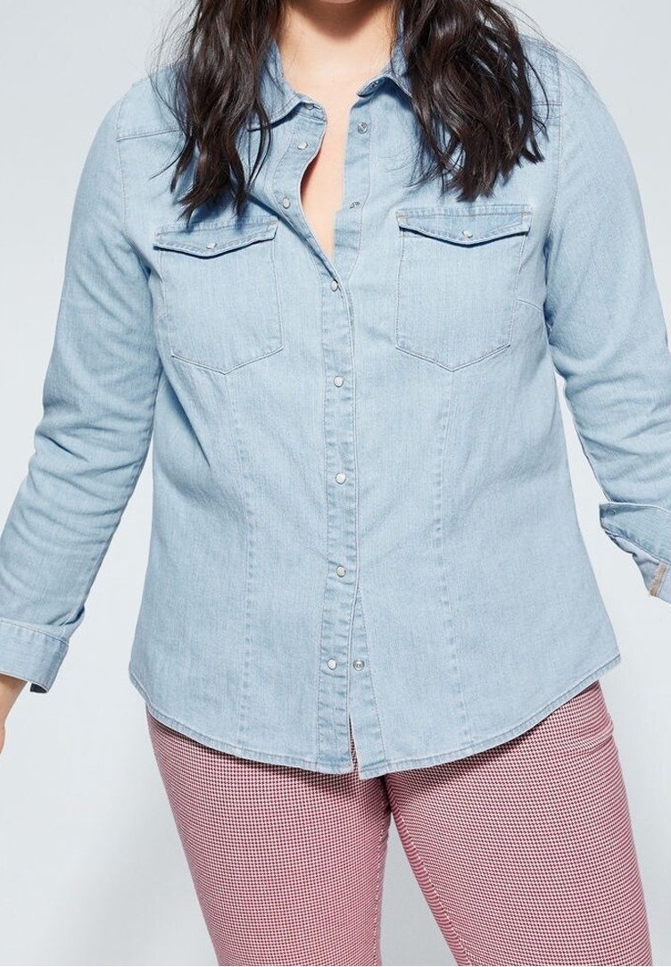 Camicia di jeans azzurro chiaro con maniche lunghe, chiusura con bottoni, due tasche sul petto e dettagli di cucitura discreti, indossata con pantaloni a quadri rosso e bianco.