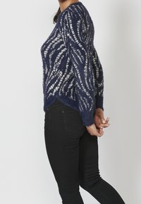 Suéter de punto azul marino con franjas onduladas blancas, acabado texturizado, cuello redondo, mangas largas y dobladillo asimétrico, combinado con jeans negros.