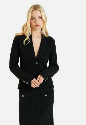 Blazer negro con un profundo escote en V, mangas largas y detalles decorativos de botones. Falda negra de canalé con bolsillos laterales y cierres de botón.