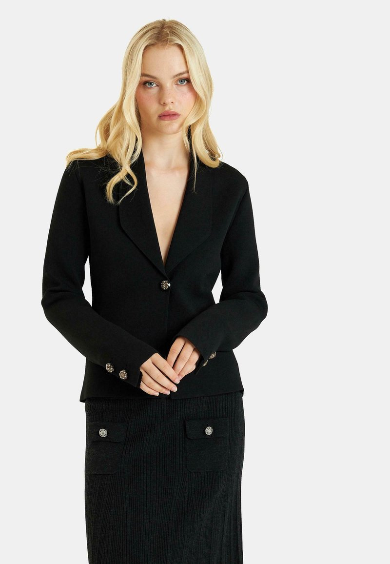 Blazer nero con profondo scollo a V, maniche lunghe e dettagli con bottoni decorativi. Gonna nera a coste con tasche laterali e chiusure a bottone.