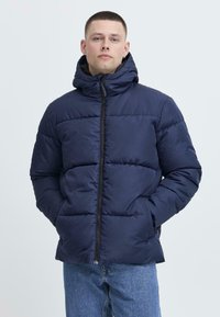 Jachetă puffer bleumarin, cu guler înalt și închidere cu fermoar, având panouri matlasate și glugă, asortată cu blugi de culoare albastru deschis.