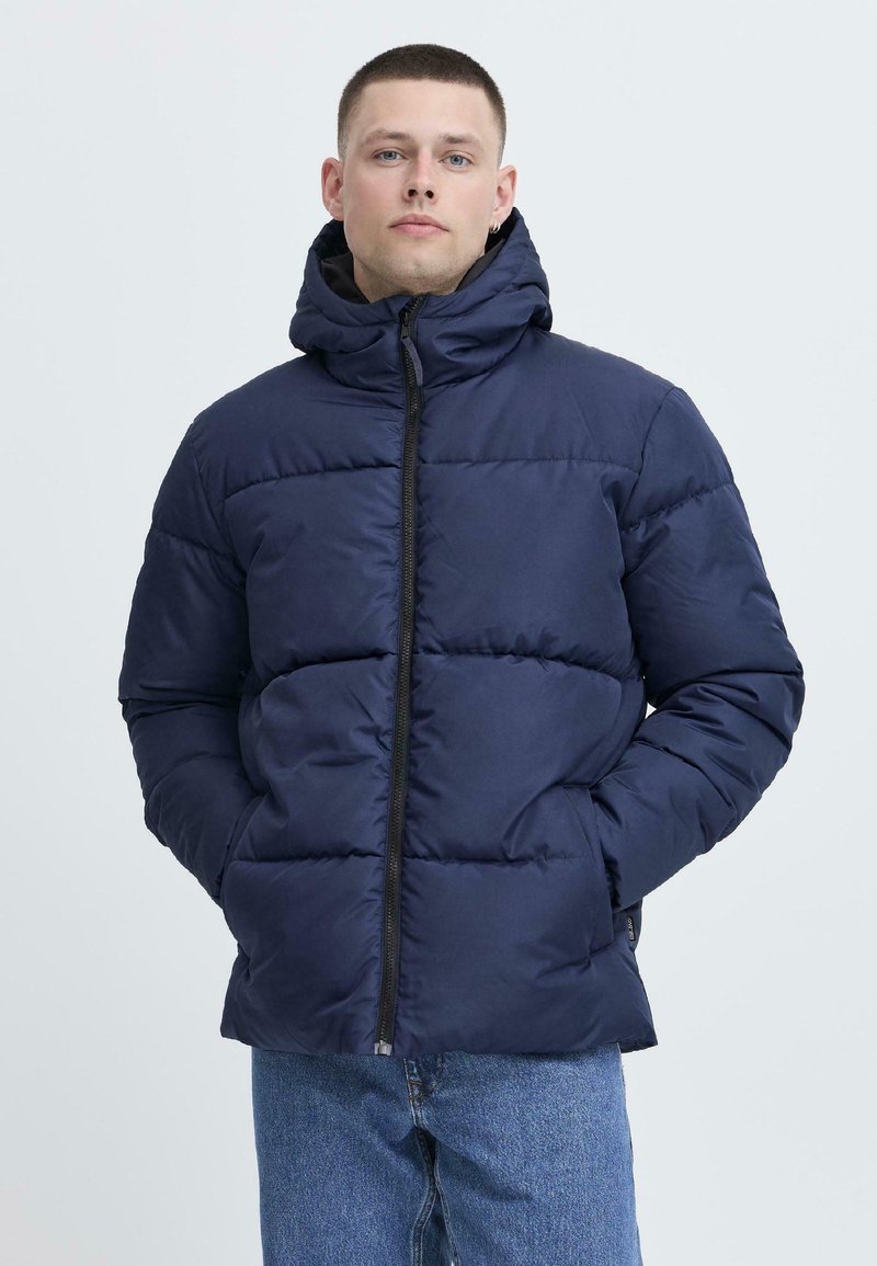 Jachetă puffer bleumarin, cu guler înalt și închidere cu fermoar, având panouri matlasate și glugă, asortată cu blugi de culoare albastru deschis.
