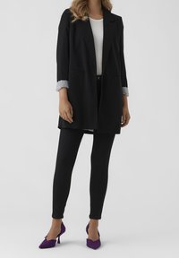 Woman wearing black blazer with rolled striped cuffs, white top, black pants, and purple textured high-heeled shoes, standing against gray background.