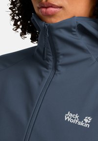 Giacca softshell blu navy con colletto alto, chiusura con zip e dettagli del logo sul petto. Il materiale sembra liscio e leggero.