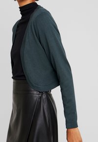 Woman wearing dark green cropped cardigan over black turtleneck with high-waisted black leather skirt against plain background.