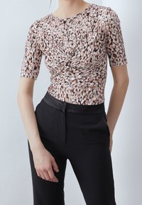 Mujer vestida con una blusa de mangas cortas de diseño torcido en marrón y blanco, combinada con pantalones negros de talle alto, sobre un fondo blanco liso.