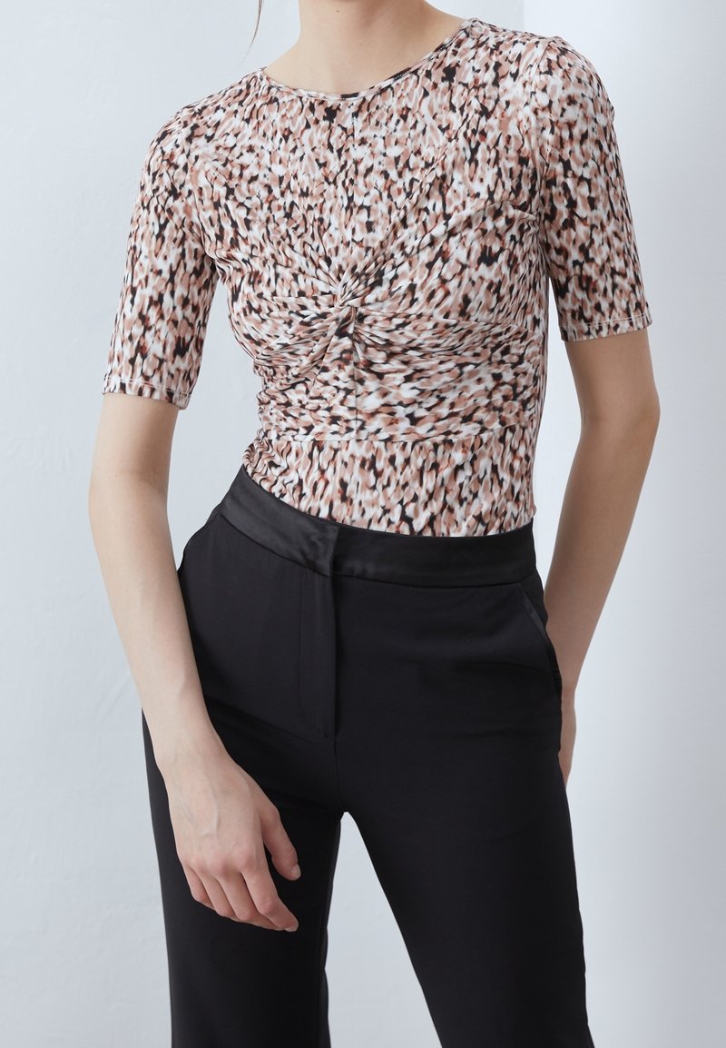 Mujer vestida con una blusa de mangas cortas de diseño torcido en marrón y blanco, combinada con pantalones negros de talle alto, sobre un fondo blanco liso.