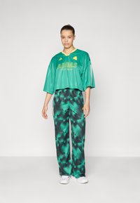 Un haut en jersey vert court avec un logo "ADIDAS" jaune, associé à un pantalon vert ample à motifs présentant un design dégradé. Des baskets blanches complètent le look.