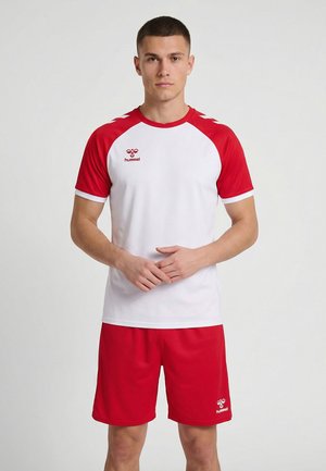 Młody mężczyzna stojący nieruchomo, ubrany w biało-czerwoną sportową koszulkę i dopasowane czerwone spodenki z logo marki na obu elementach.