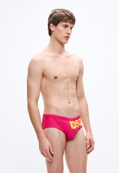 Rožinės maudymosi kelnaitės su geltonu "DSQ" logotipu priekyje. Aptemptas dizainas, minkšta tekstūra, šoninis vaizdas, atskleidžiantis koją ir juosmenį.