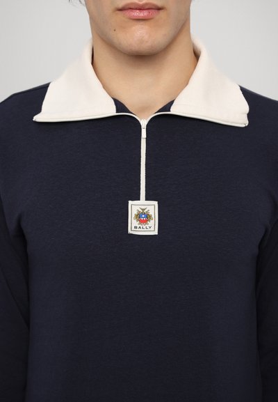 Navy zilu krekls ar garām piedurknēm un krēma krāsas rāvējslēdzēja apkakli, kurā ir centrēts Bally logotipa uzlīme ar dekoratīviem elementiem. Mīksta auduma tekstūra.