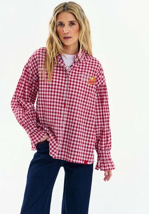 Kobieta o długich blond włosach, ubrana w czerwoną i białą koszulę w kratę z falbankami na mankietach oraz ciemnoniebieskie dżinsy, stojąca na tle białego tła.