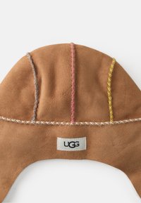 Ruskea mokkanahkahattu, jossa on eriväriset kirjonnat sinisen, vaaleanpunaisen ja keltaisen sävyissä. Hatussa on UGG-labeli pohjassa. Pehmeä tekstuuri ja pyöreä muoto.