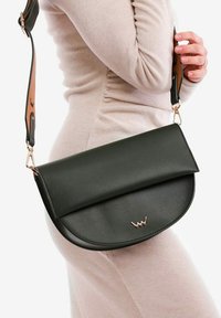 Zelená kožená crossbody taška s kulatým klopou, vybavená nastavitelným popruhem s béžovými akcenty a malým zlatým logem. Hladká textura.