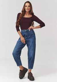 Bordowy top z długim rękawem w połączeniu z niebieskimi dżinsami o wysokim stanie i luźnym kroju. Modelka nosi czarne trampki na platformie. Proste tło.
