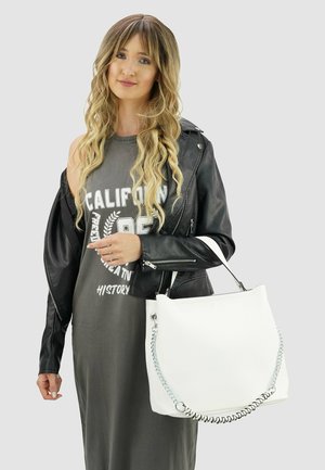 Jonge vrouw met lang golvend blond haar draagt een leren jas over een grijze jurk met tekst, en houdt een grote witte handtas met kettingdetail vast.