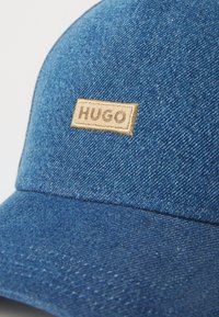 Σκούρο μπλε τζιν καπέλο baseball με υφή, που διαθέτει μία μπεζ ετικέτα από ύφασμα με την επιγραφή "HUGO". Κυρτή γείσος και ραμμένα λεπτομέρειες.