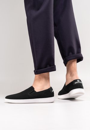 Benenden met donkergekleurde opgerolde broeken en zwarte instapsneakers met witte zolen, met het merklabel "GIESSWEIN" op het hielbandje.