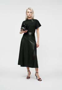 Femme blonde portant un haut noir à manches courtes, une jupe midi en cuir noir avec fente, des sandales à talons hauts noires, tenant une pochette texturée noire.