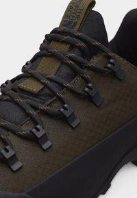 Κοντινό πλάνο παπουτσιού πεζοπορίας σε ελιά πράσινο με κορδόνια με μαύρο σχέδιο, μαύρους γάντζους κορδονιών και ταμπελάκι γλώσσας με λογότυπο, που διαθέτει υφή στο άνω μέρος και παχιά σόλα.