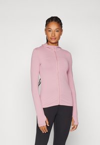 Ljusrosa zip-up hoodie med en texturerad tygupplevelse, långa ärmar och tumhål, tillsammans med svarta leggings.